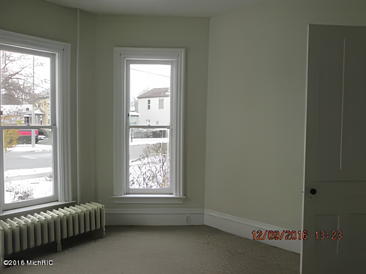 An empty room with two large windows and a radiator beneath, showing a snowy exterior scene.