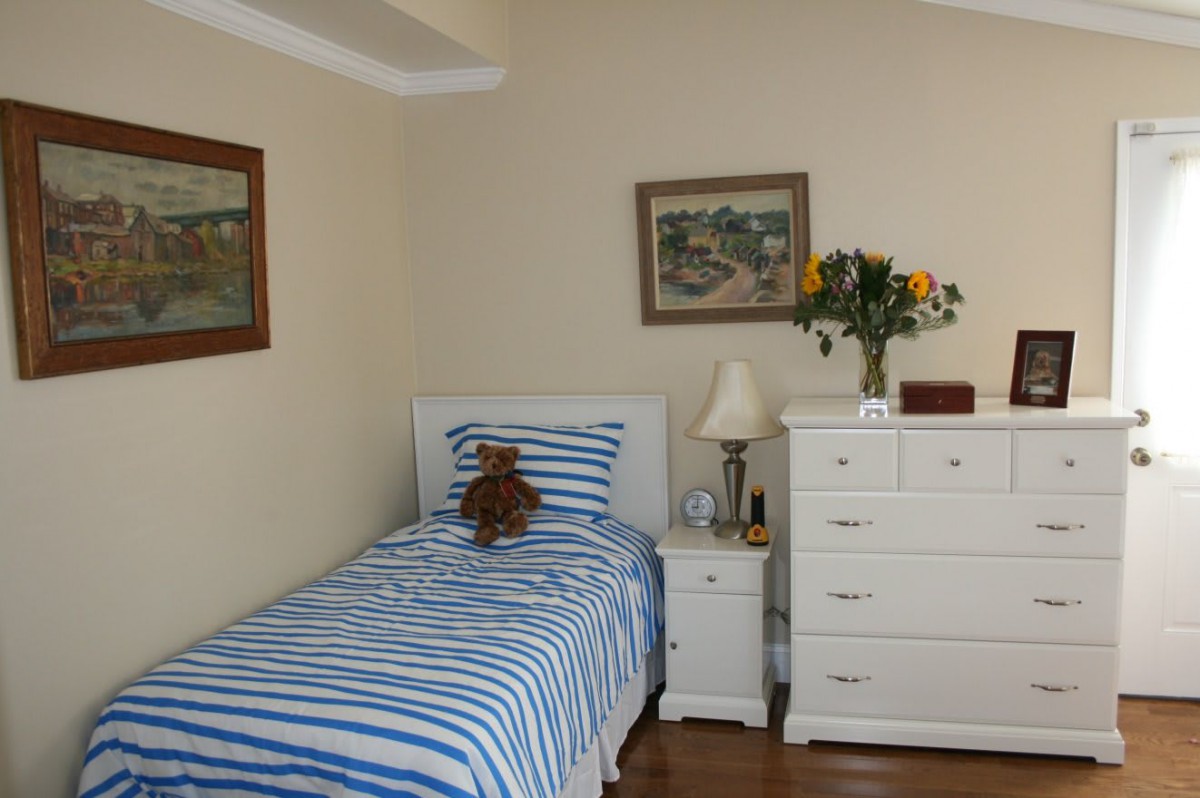 A cozy bedroom with a single bed featuring striped bedding, and a dresser topped with flowers and a lamp.