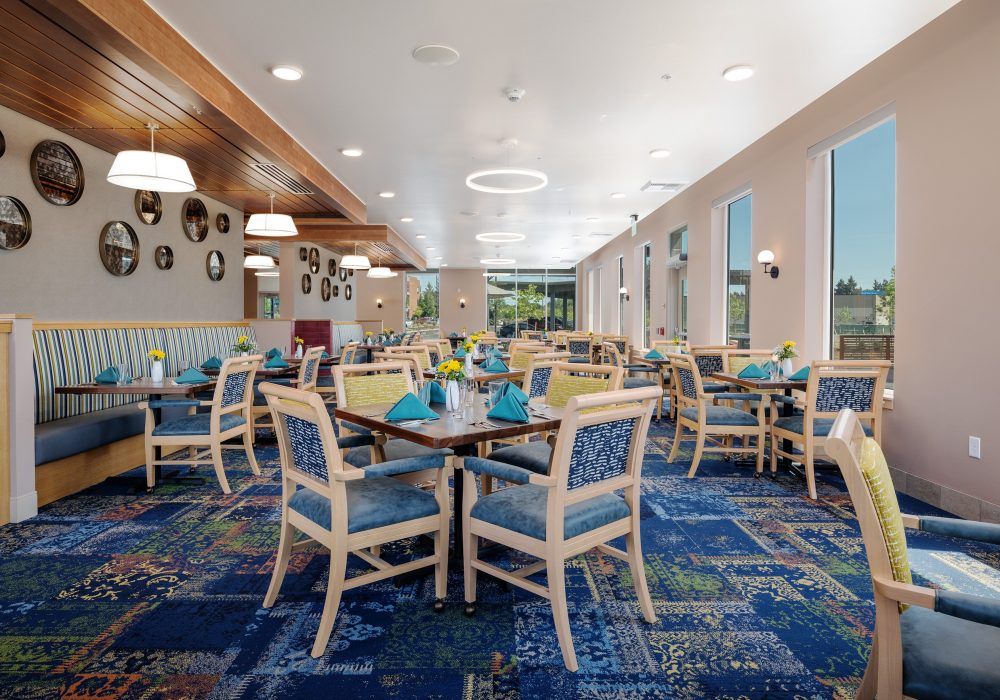 A modern dining room with neatly arranged tables and chairs, featuring a patterned carpet and ample natural light.