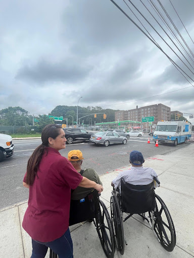 A caregiver assisting two individuals in wheelchairs on a busy city street under a cloudy sky.