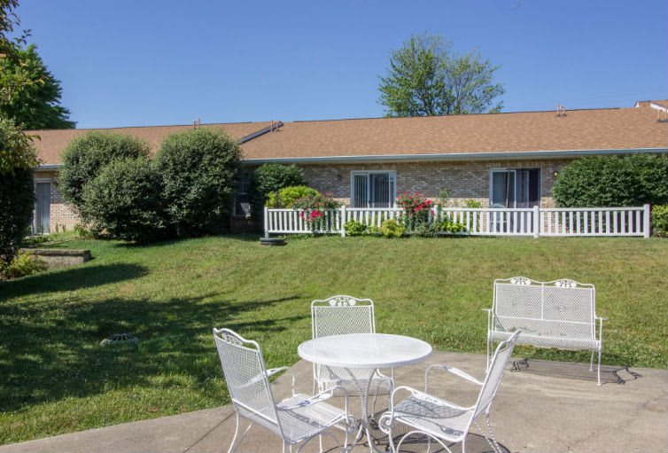 A sunny outdoor patio with a white table and chairs, overlooking a well-maintained garden and lawn in front of a house.
