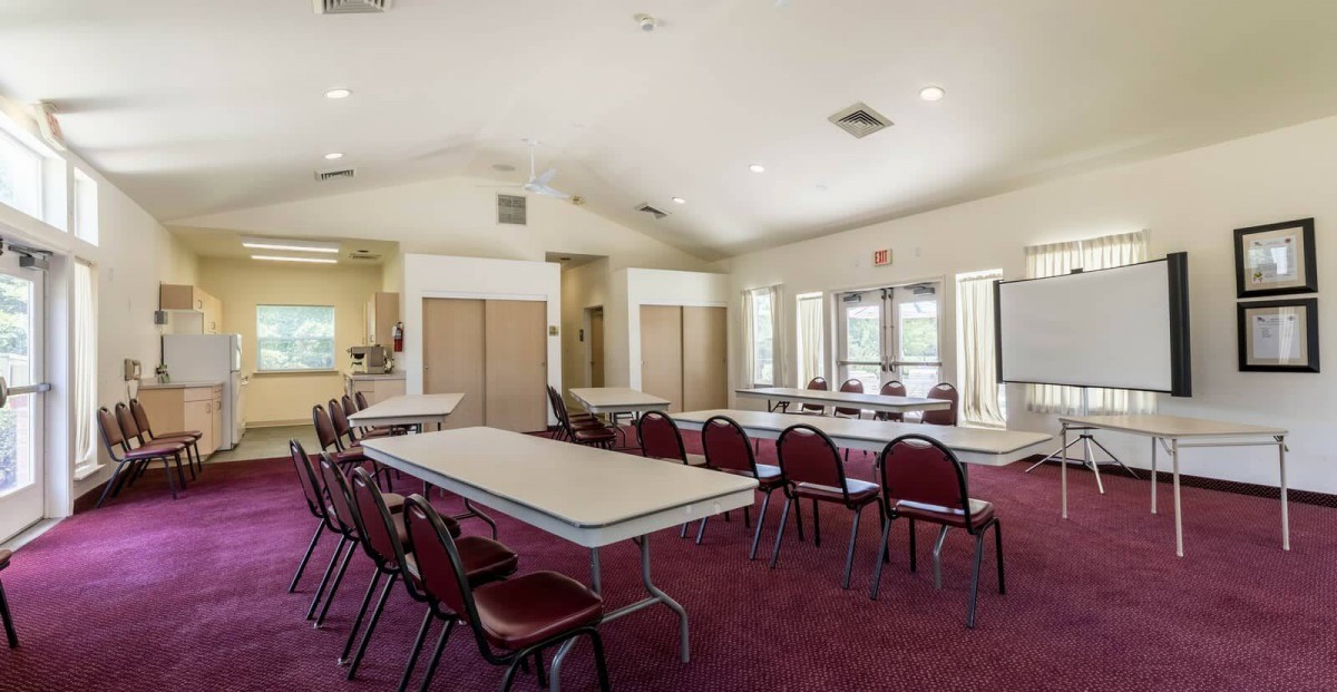 A spacious conference room with tables and chairs arranged on a carpeted floor, featuring large windows for natural light.