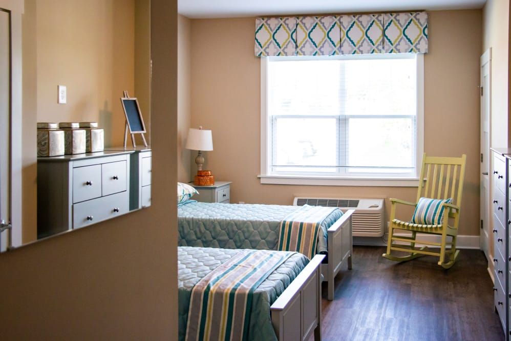 A well-lit hospital room with twin beds, featuring a lamp on the nightstand and a rocking chair by the window.