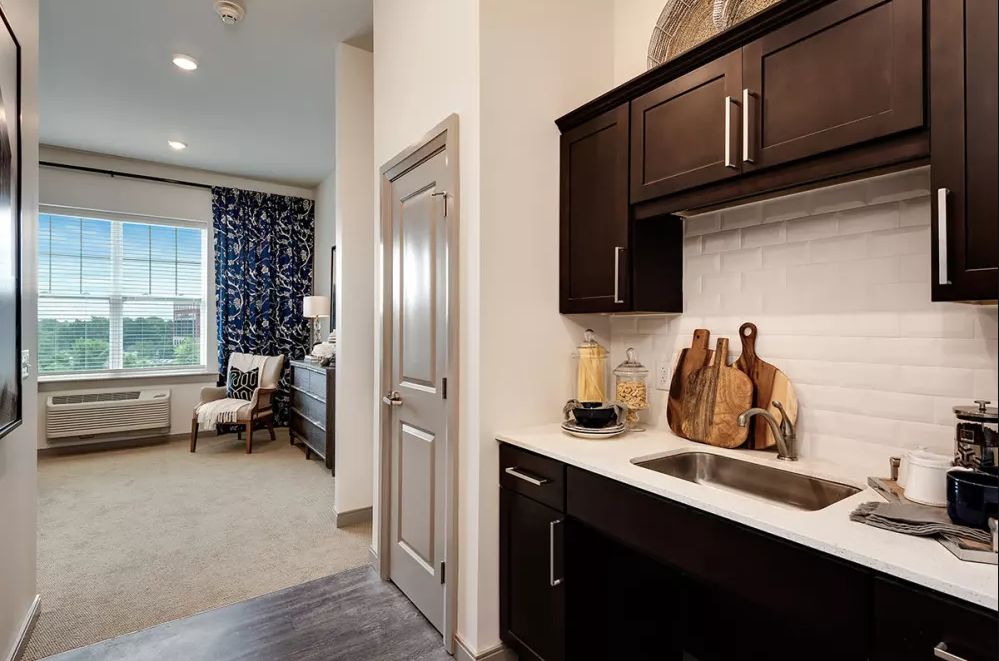 A modern kitchenette with dark cabinets opens into a cozy, well-decorated bedroom.
