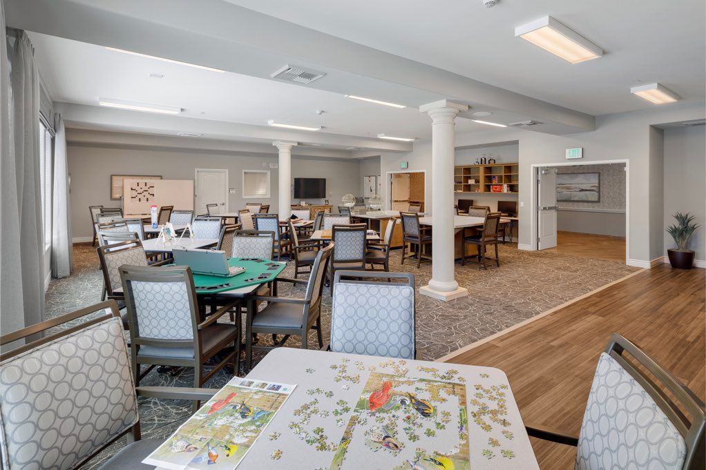 A well-lit community dining area with tables and chairs, featuring a puzzle in progress.