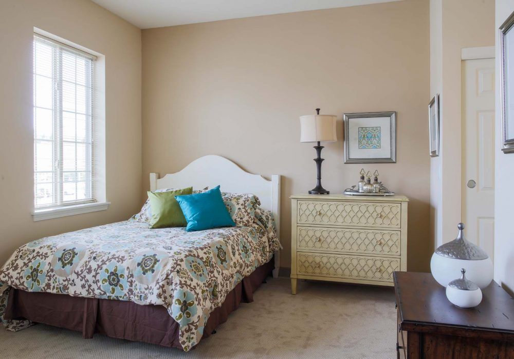 A cozy bedroom with a bed, patterned bedding, and a decorative lamp on a chest of drawers.