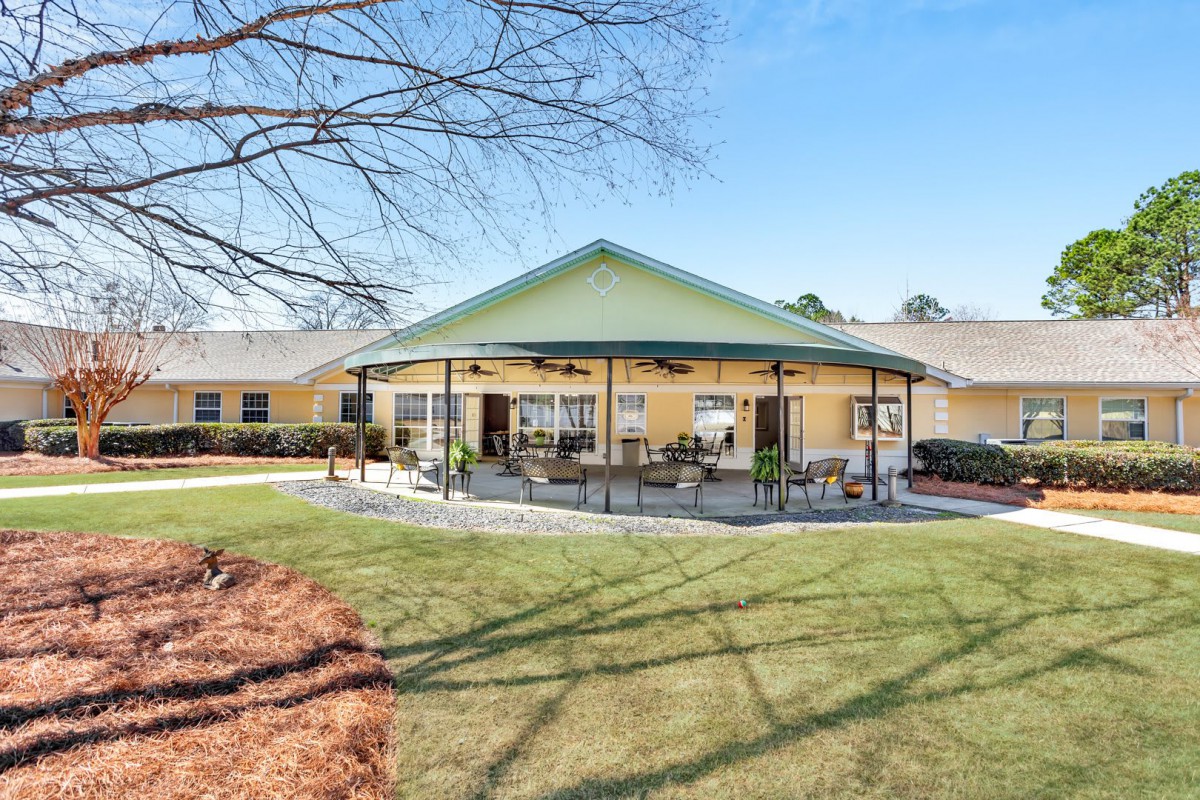 A view of a building with a patio and outdoor seating area, surrounded by a grassy yard and trees.