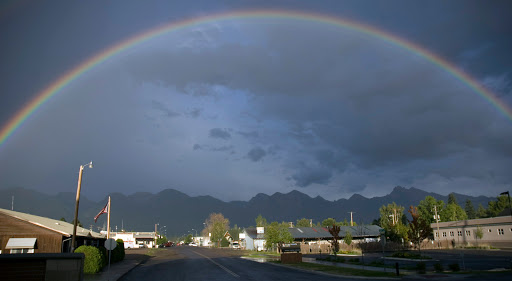 A stunning rainbow arches over a quiet road, with mountains in the background and dark clouds hinting at recent rain.