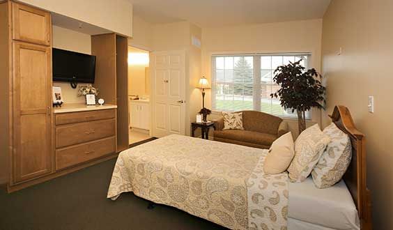 A cozy bedroom with a bed, couch, and television, featuring soft lighting and a large window.