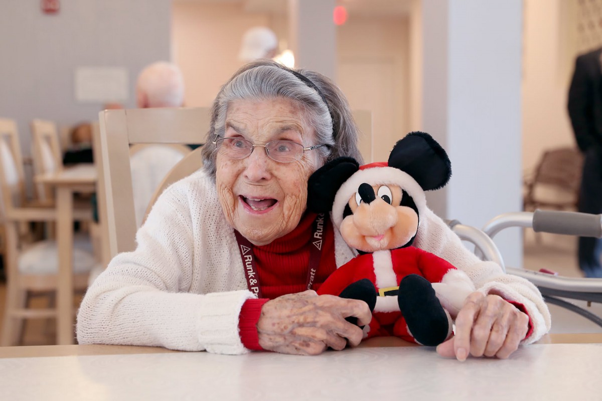 An elderly woman smiles warmly while holding a Mickey Mouse plush toy dressed in a holiday outfit indoors.