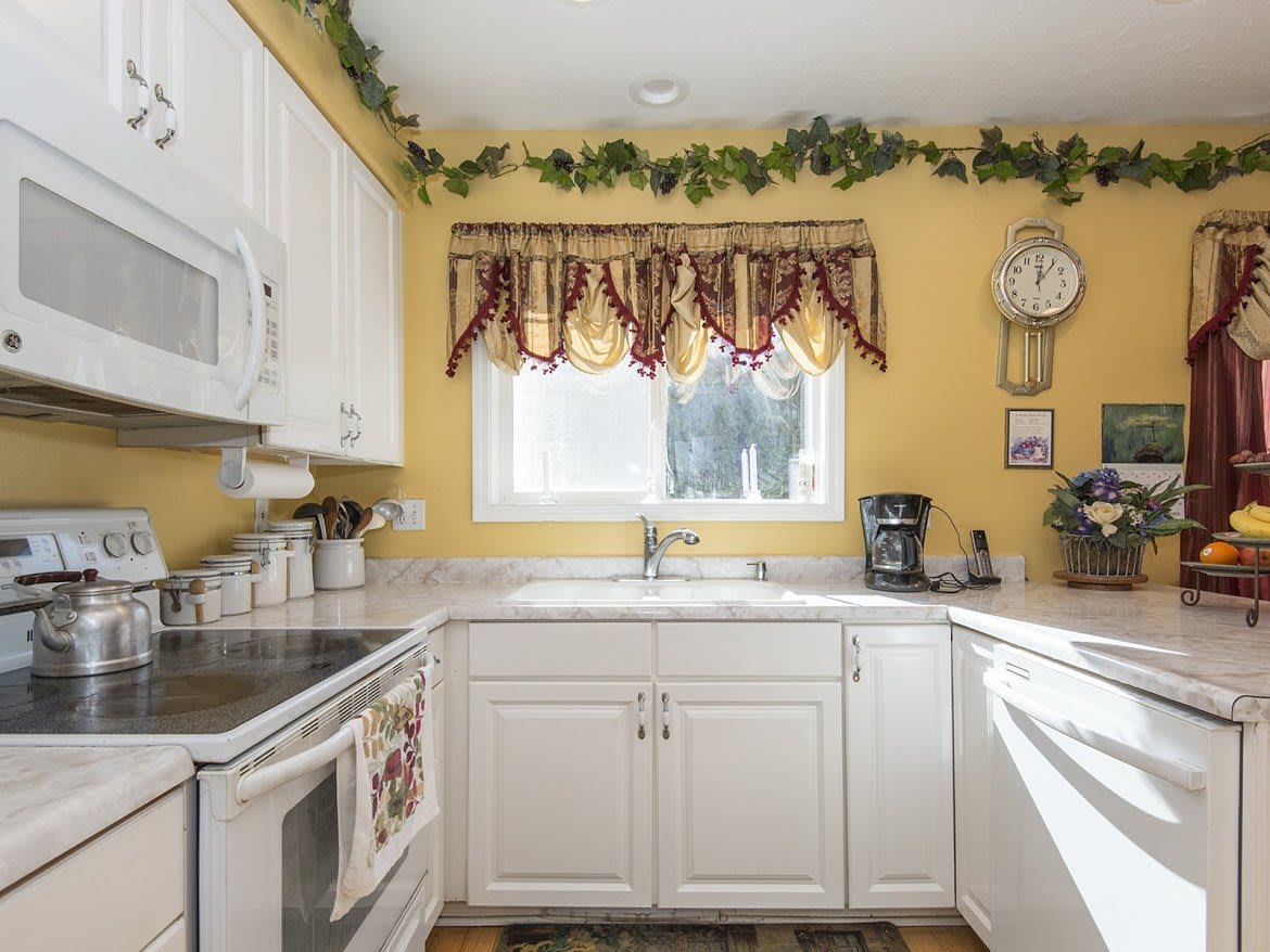 A cozy kitchen with white cabinets, yellow walls, and a window adorned with red and gold curtains.