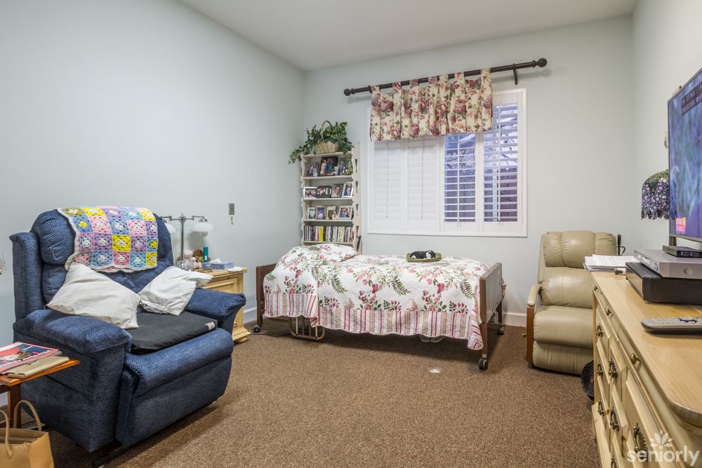 A cozy bedroom featuring a single bed, a blue armchair, and a beige recliner with a bookshelf filled with books.