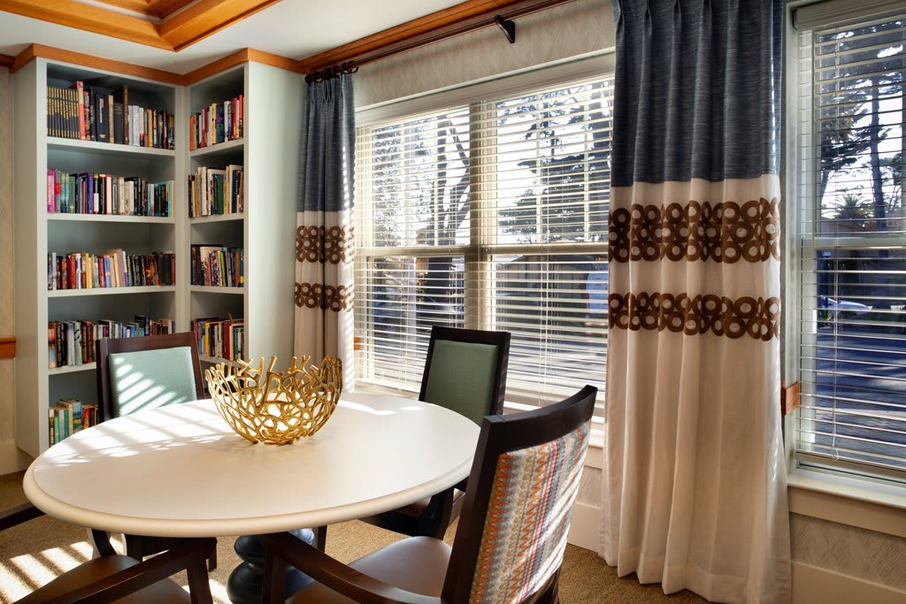 A cozy dining room with a round table, chairs, large window with curtains, and a bookshelf filled with books.