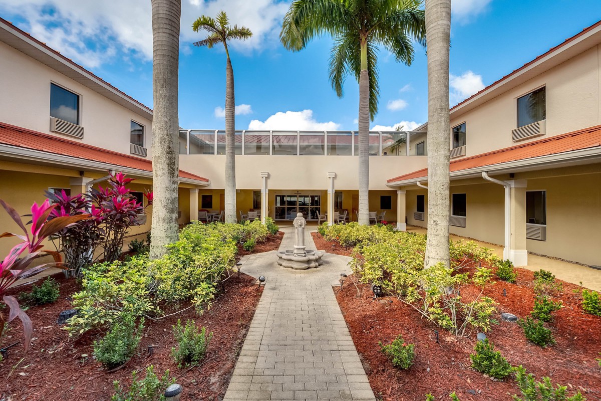 A serene courtyard with a central pathway, lined with palm trees and a fountain, leading to a building under a clear blue sky.