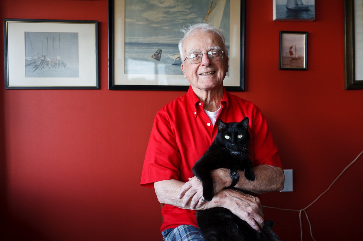 An elderly man in a red shirt smiling and holding a black cat indoors.