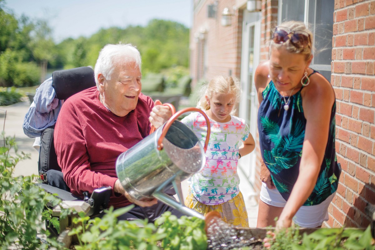 An elderly man in a wheelchair watering plants with the help of a woman and a young girl outdoors.
