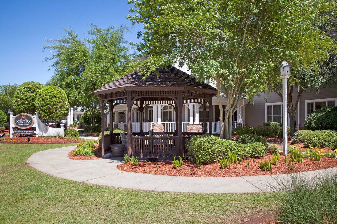 A charming wooden gazebo surrounded by well-maintained landscaping and a paved pathway under a clear blue sky.