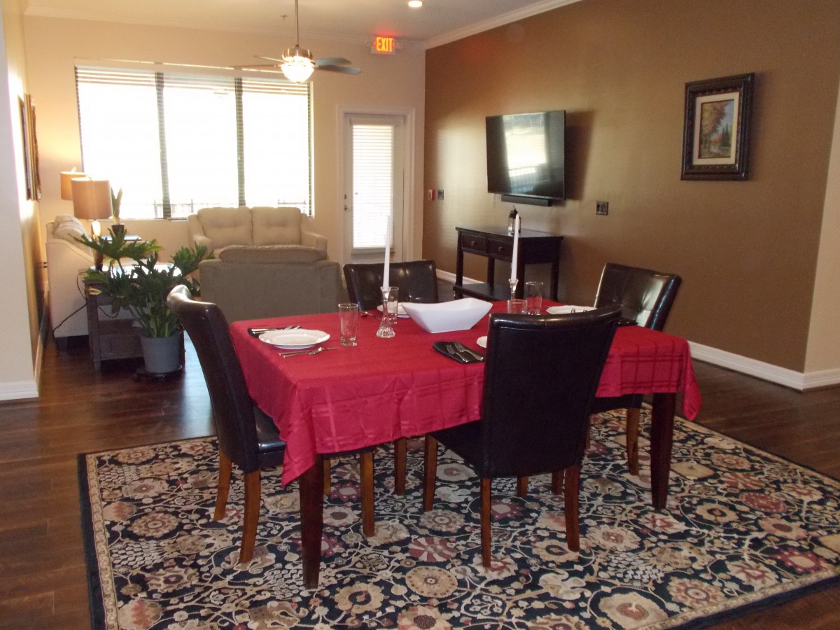A dining area with a red tablecloth and black chairs, adjacent to a living room featuring a television on the wall.