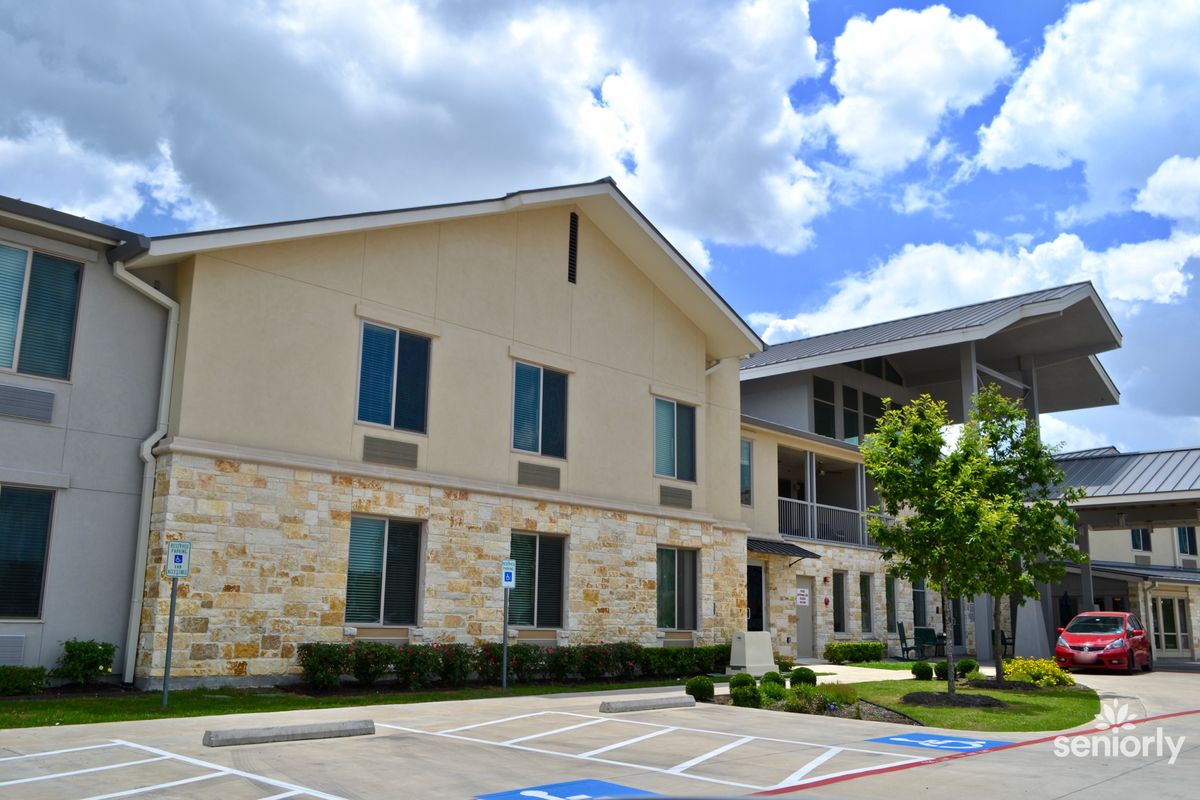 A residential building with a stone facade and several windows, set against a bright blue sky with clouds. Visible in the foreground is a parking lot with designated spaces.