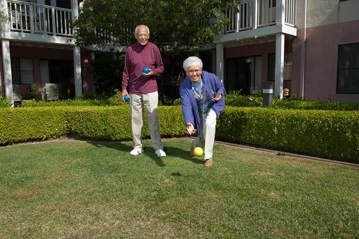 An elderly couple enjoys playing bocce ball outdoors in a garden setting.