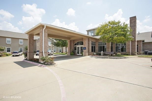 A building entrance with a covered driveway and surrounding trees on a sunny day.