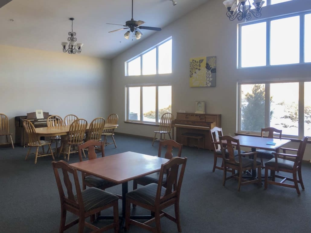 A spacious dining area with large windows, featuring multiple tables and chairs, illuminated by natural light and ceiling fixtures.