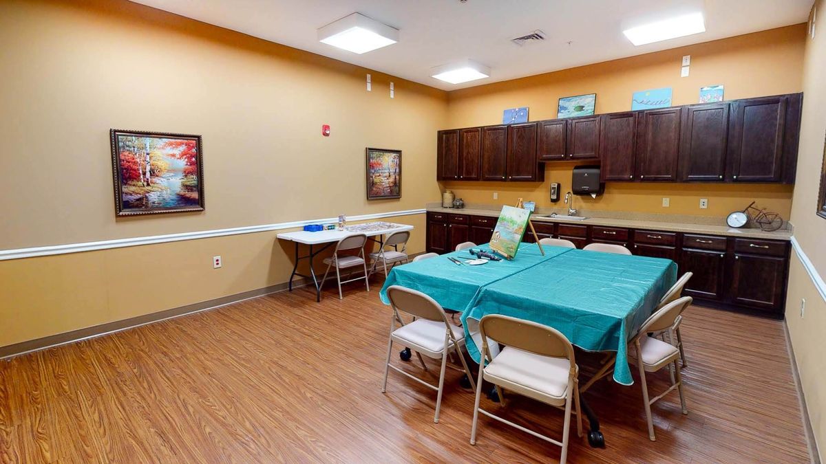 A multi-purpose room with tables and chairs, featuring kitchen cabinets and wall art.