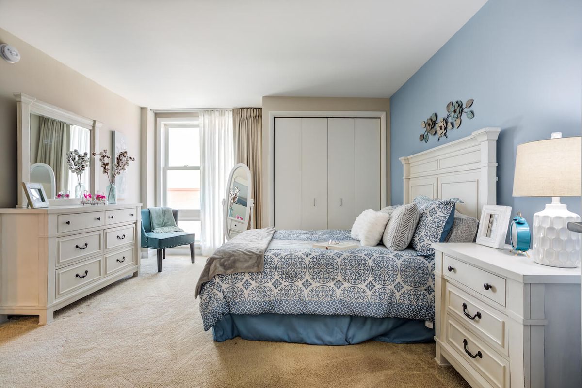 A cozy bedroom with blue walls, featuring a bed, dresser, and chair, bathed in natural light.