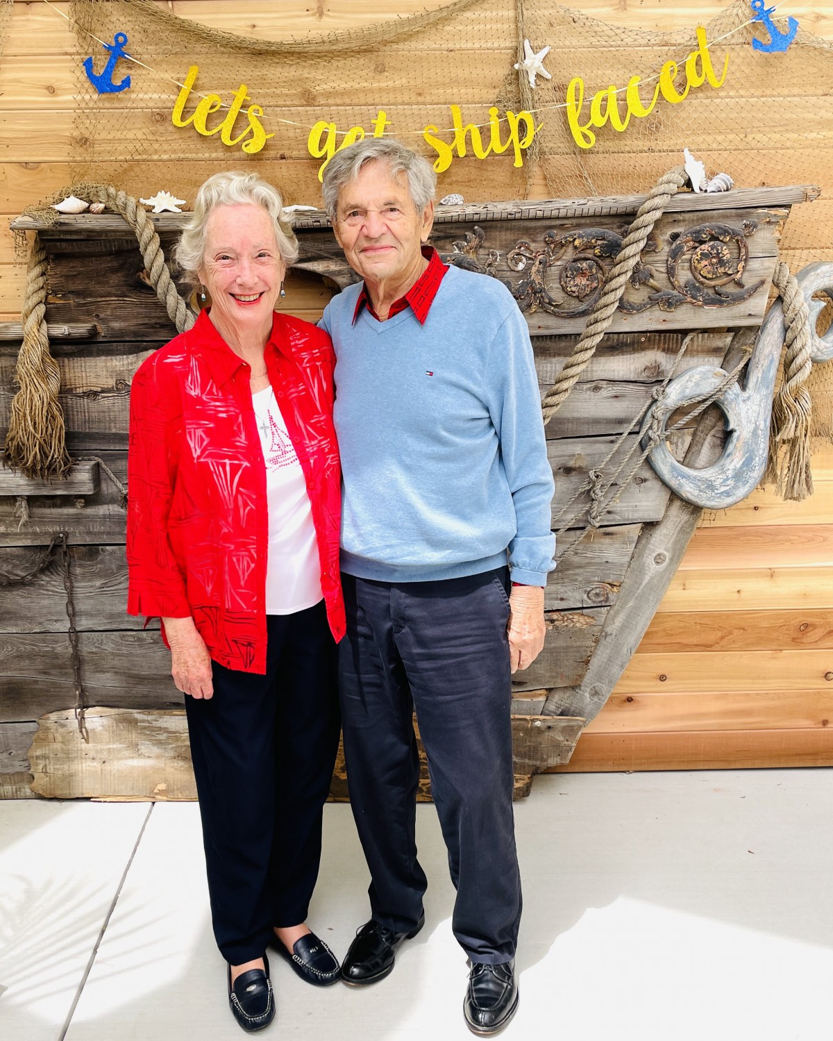 An elderly couple smiling together in front of a decorative wooden backdrop with ropes.