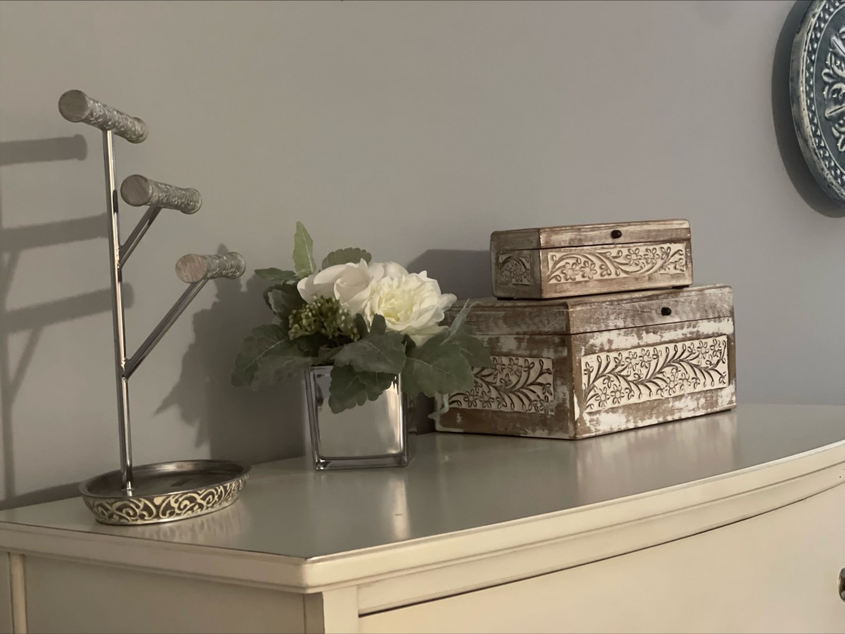 A decorative setup featuring a bouquet of white flowers in a vase, ornate boxes, and a jewelry holder on a dresser.
