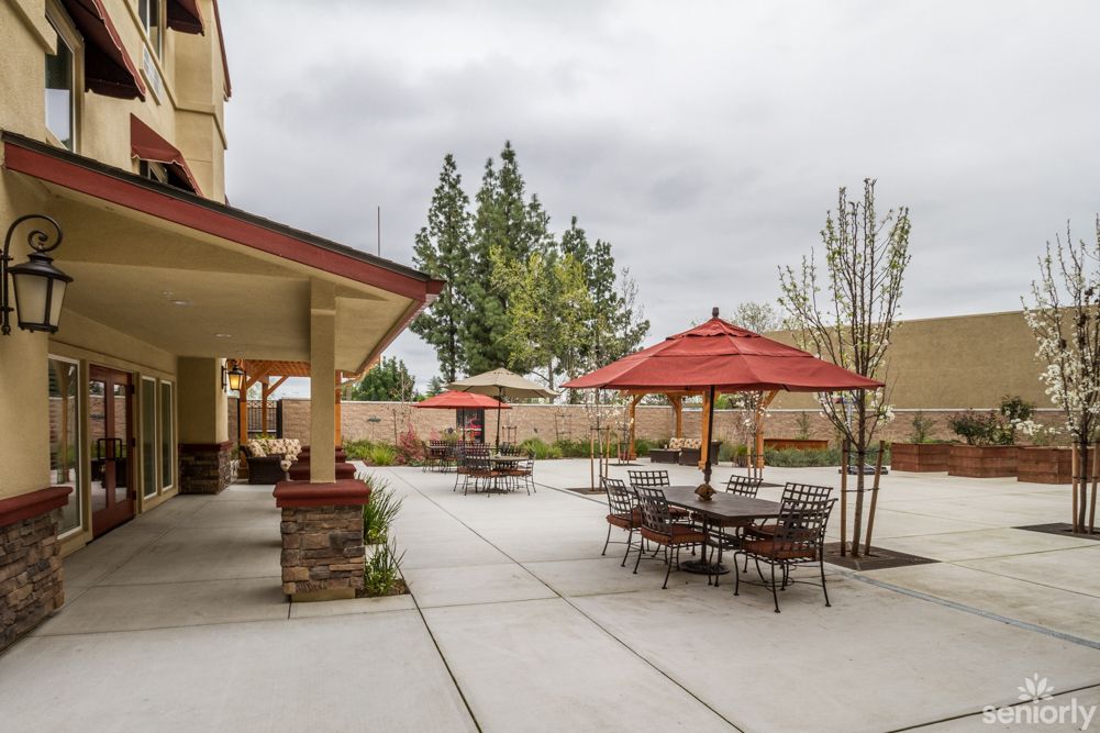 A paved outdoor patio area with tables, chairs, and red umbrellas set against a backdrop of trees under a cloudy sky.