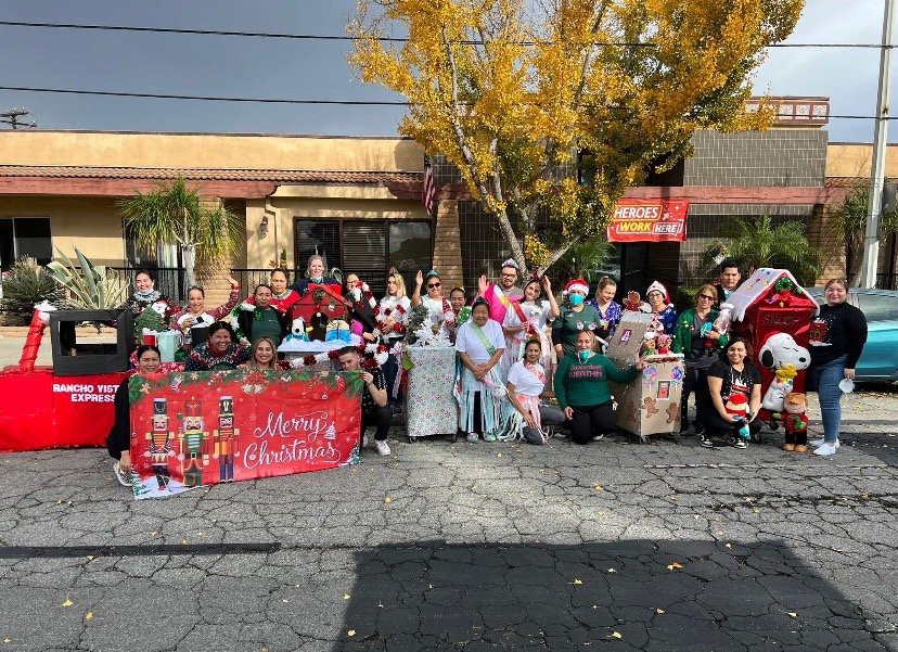 A festive group gathers outdoors for a Christmas celebration, displaying a colorful banner and wearing holiday-themed costumes.