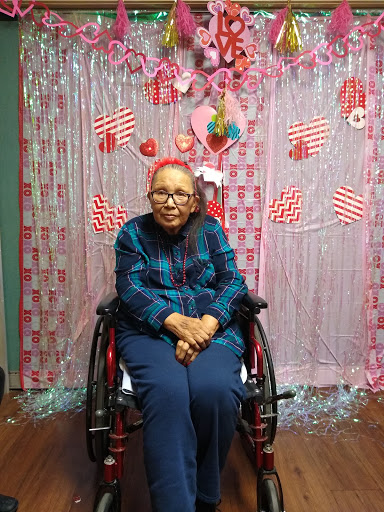 An elderly woman in a wheelchair sits against a colorful Valentine's-themed backdrop.