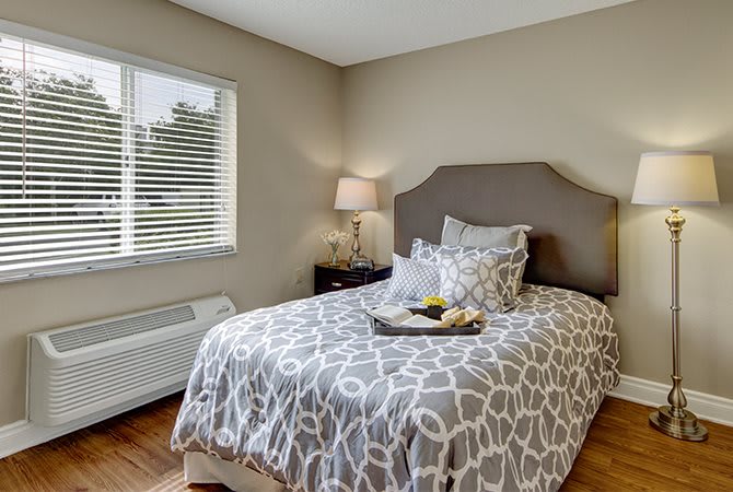 A cozy bedroom featuring a bed with patterned bedding, a bedside table with a lamp, and a window with blinds.