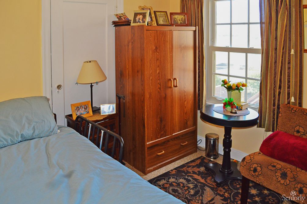 A cozy bedroom with a wooden wardrobe, side table with a lamp, and a round table near the window.