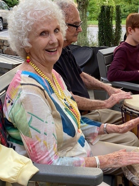 An elderly woman smiling while participating in an outdoor drumming session with others.