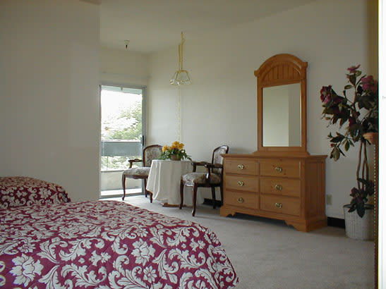 A cozy bedroom featuring a wooden dresser with a mirror, a bed with a patterned bedspread, and a small table with chairs and flowers.