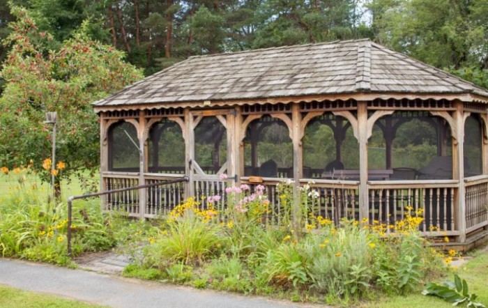 A wooden gazebo surrounded by lush garden greenery and flowers.