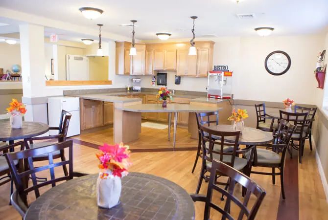 A spacious kitchen and dining area with wooden flooring, featuring several tables and chairs, pendant lights, and decorative flowers.