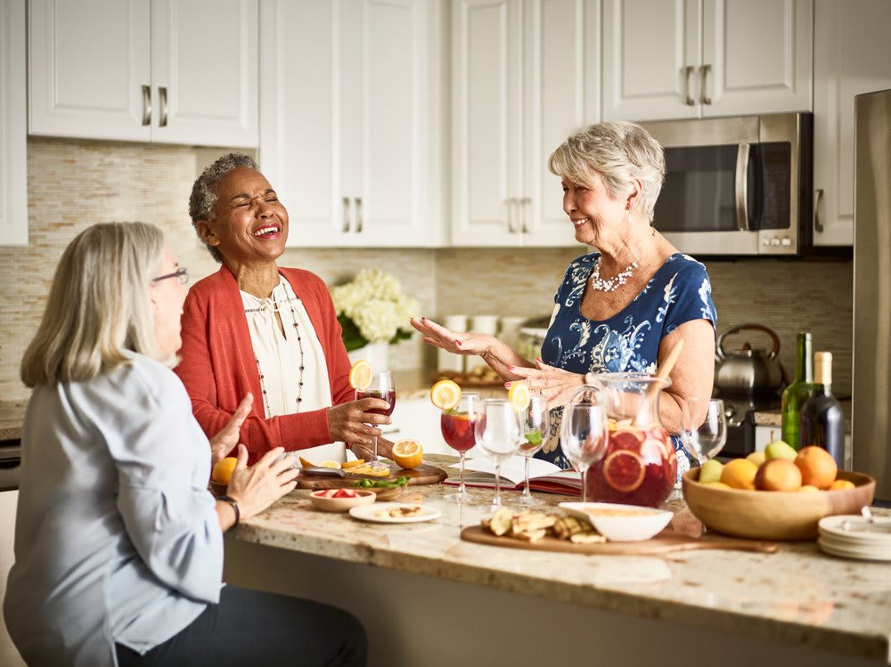 A group of people enjoying conversation and laughter in a kitchen while preparing food and drinks.