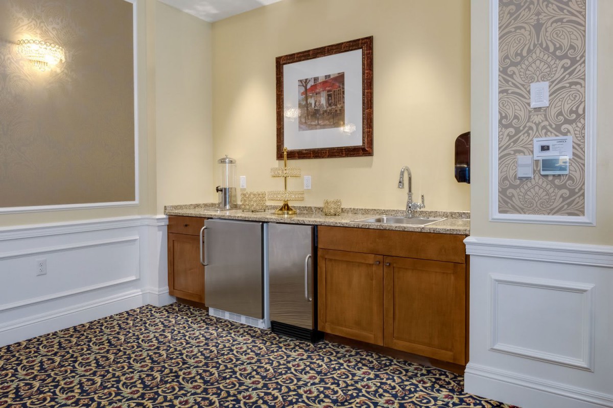 A small kitchenette with wooden cabinets, a sink, and a mini fridge, decorated with framed artwork above the countertop.