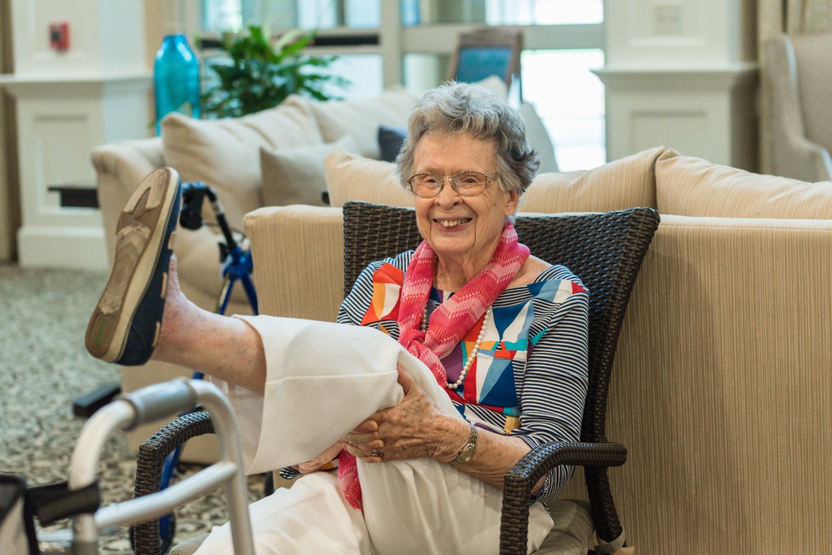 An elderly woman with glasses and a colorful scarf smiles while sitting in a wheelchair indoors.