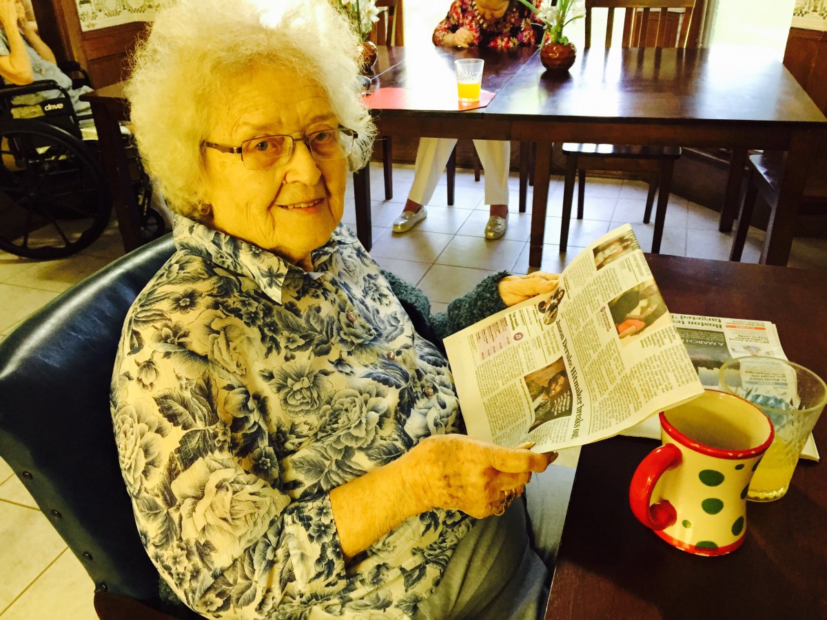 An elderly woman smiling as she reads a newspaper while sitting indoors with a mug on the table.