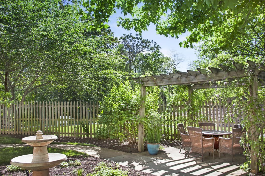 A tranquil garden scene with a wooden pergola, surrounded by lush greenery and furnished with patio seating.