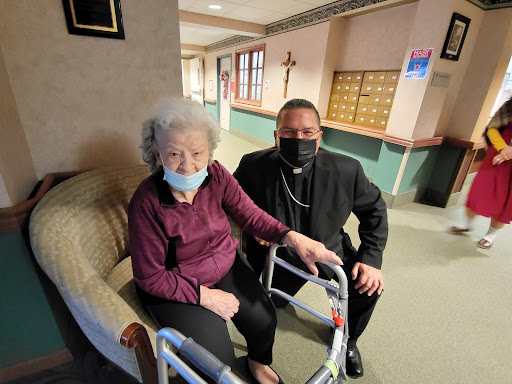 An elderly woman and a man in formal attire pose together indoors, both wearing masks.