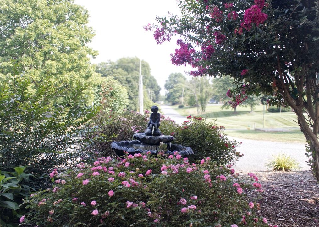 A garden scene with a small statue surrounded by blooming flowers and lush trees.