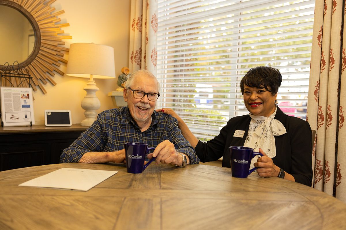 An elderly man and a woman smiling while sitting at a table with coffee mugs.