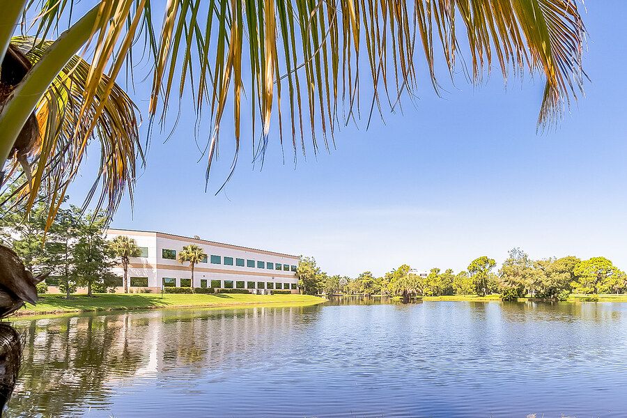 A serene view of a building by a lake surrounded by palm trees under a clear blue sky.