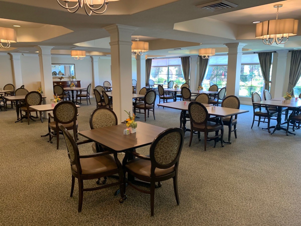 An empty dining room with neatly arranged tables and chairs, featuring large windows and elegant chandeliers.