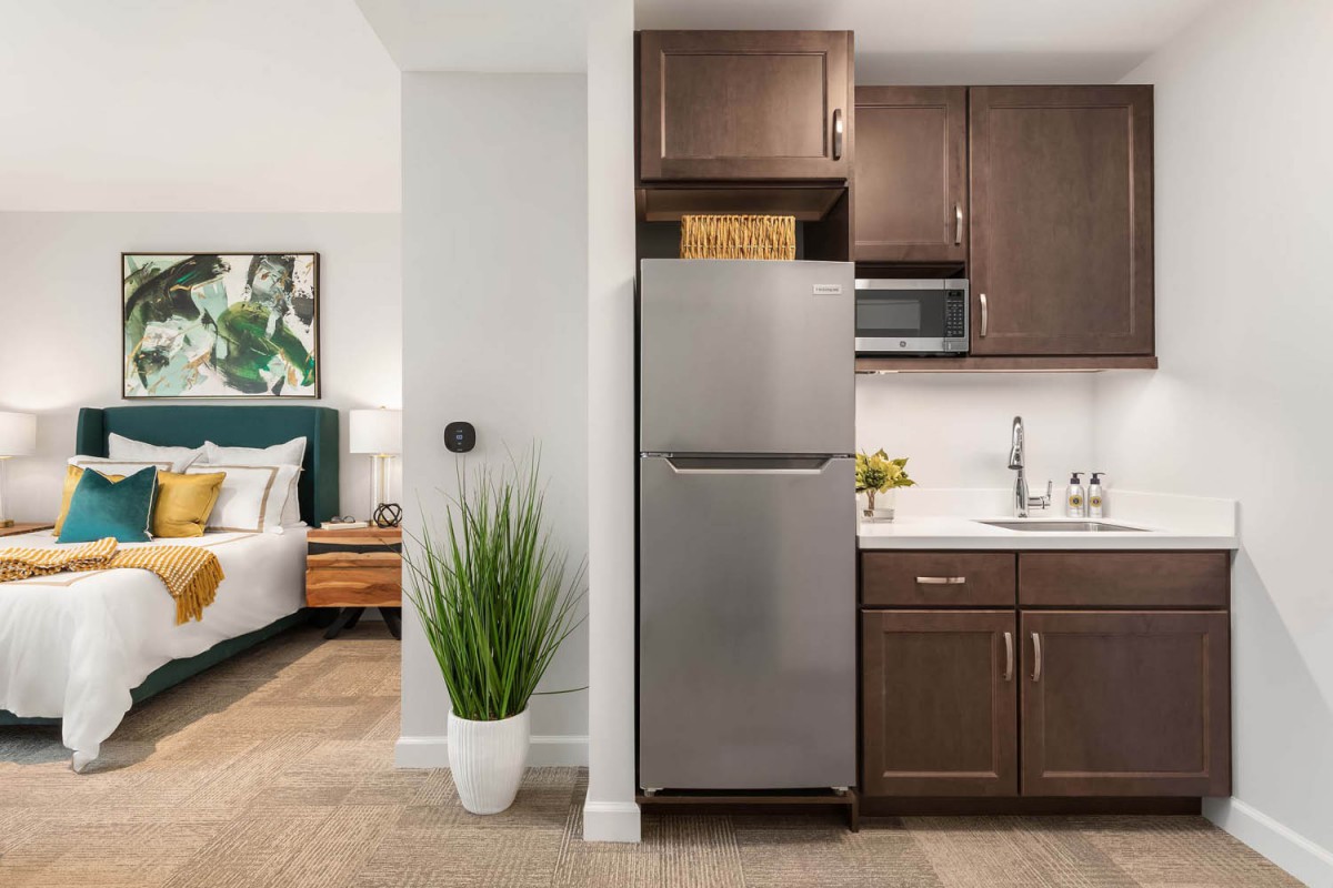 A modern apartment with an open-plan kitchen and bedroom, featuring sleek cabinetry and a stainless-steel refrigerator.